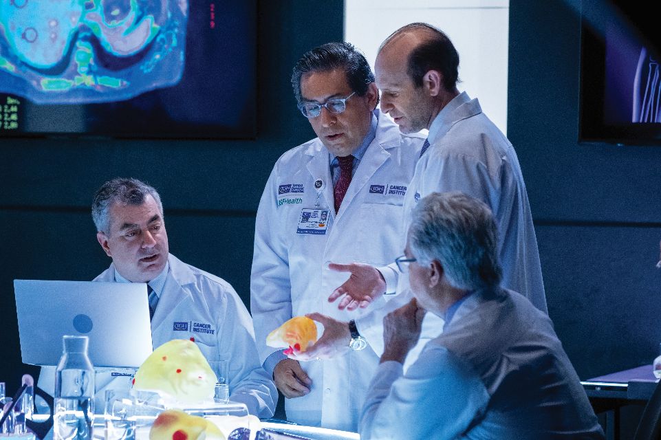 Four doctors seated and standing around a table in a darkened high-tech imaging room looking at 3-D printed models.