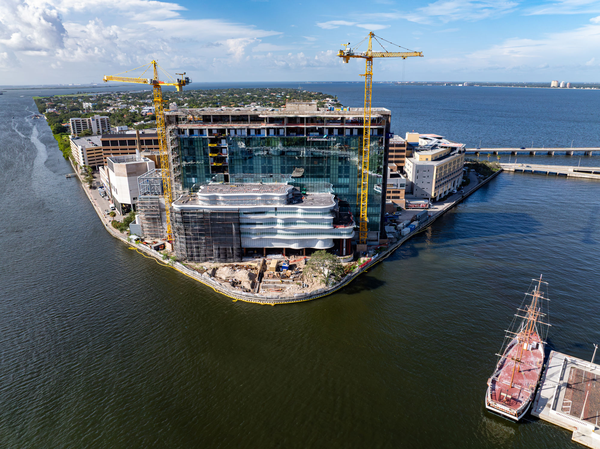 Aerial view of Tampa General Hospital’s Taneja Tower under construction on the waterfront, with two cranes, glass exterior panels, and Tampa Bay in the background.