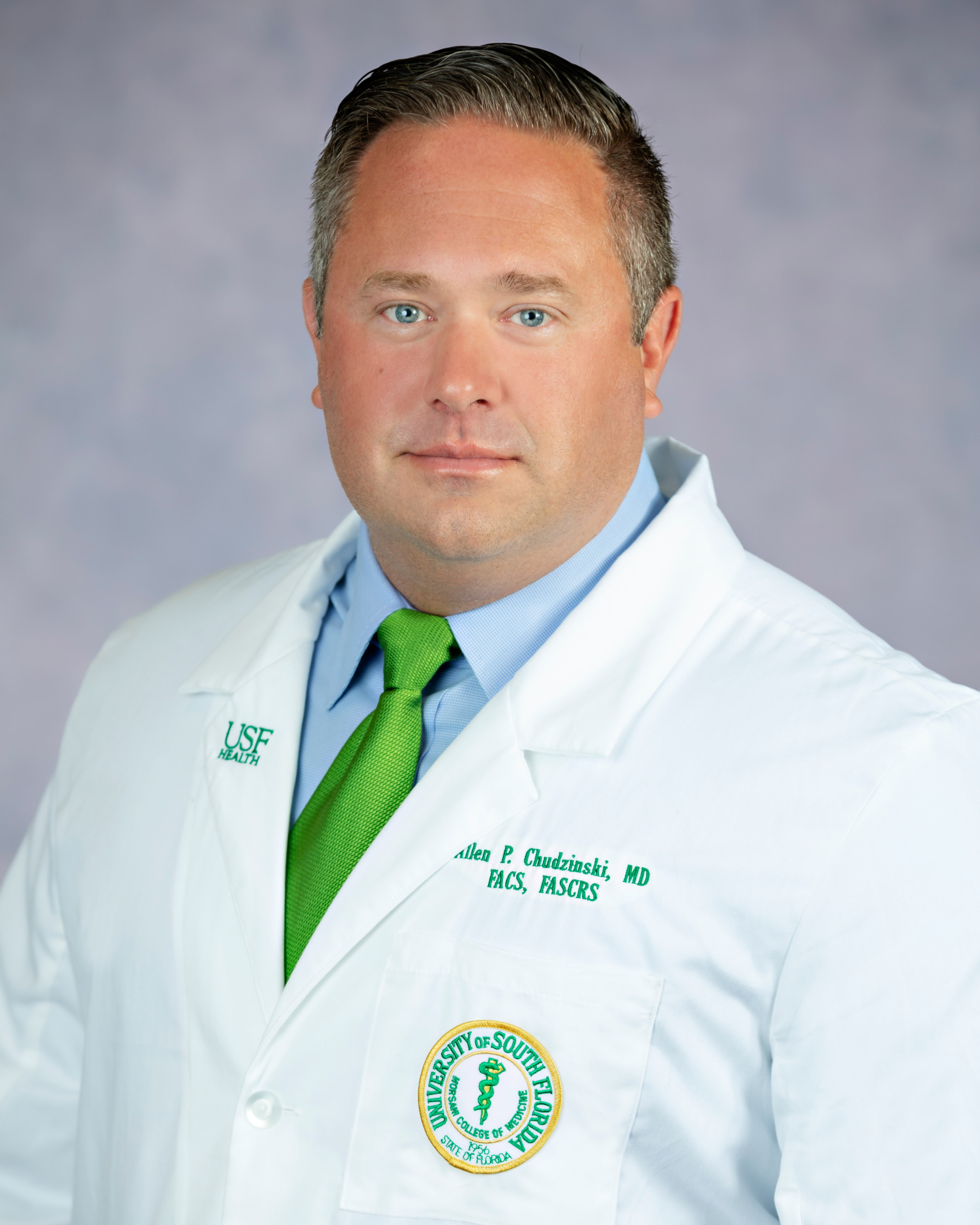 Headshot of a healthcare provider wearing a white coat with a USF Health logo, blue shirt, and bright green tie, facing forward against a soft gray background; an embroidered medical school patch is visible on the coat.
