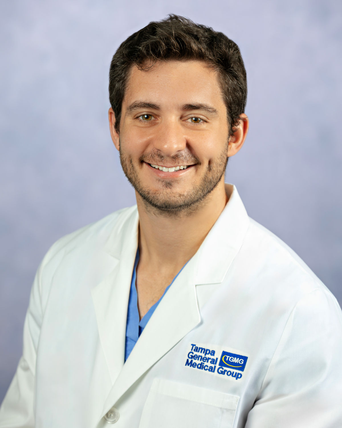 Picture of Dr. Matias Czerwonko, Thoracic Oncologist at TGH Cancer Institute, wearing a white medical lab coat with the Tampa General Medical Group logo embroidered on the chest, over light blue scrubs, posed against a soft, light-colored studio background.