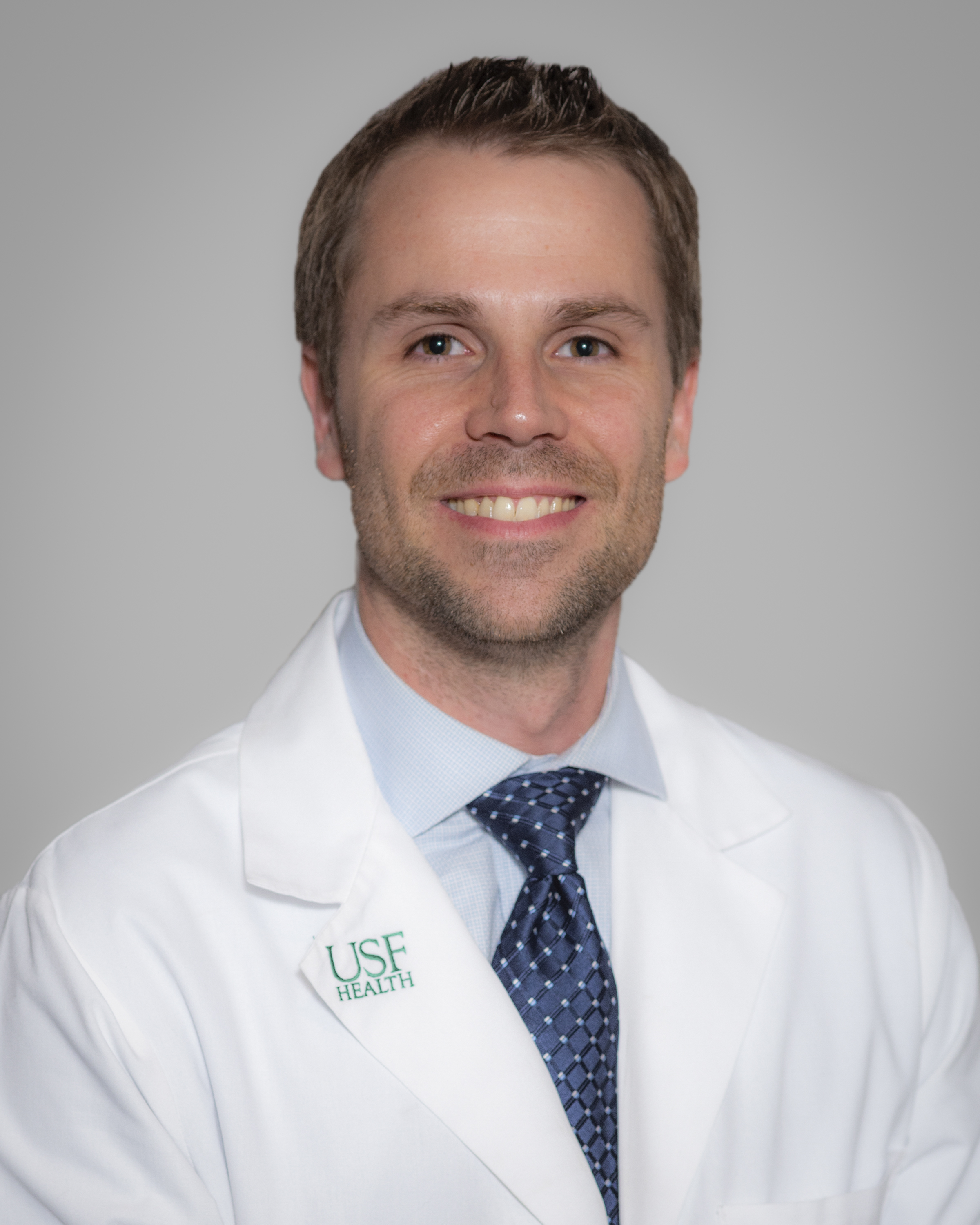 Headshot of a healthcare provider wearing a white coat with a USF Health logo, light blue shirt, and patterned tie, facing forward against a neutral gray background.