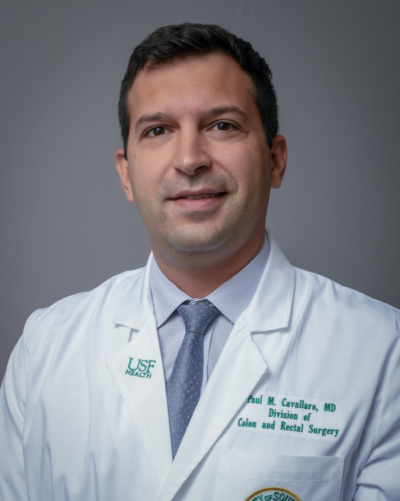 Headshot of a healthcare provider in a white coat with a USF Health logo, light dress shirt, and gray tie, facing forward against a gray background; embroidered text on coat indicates a colorectal surgery division.