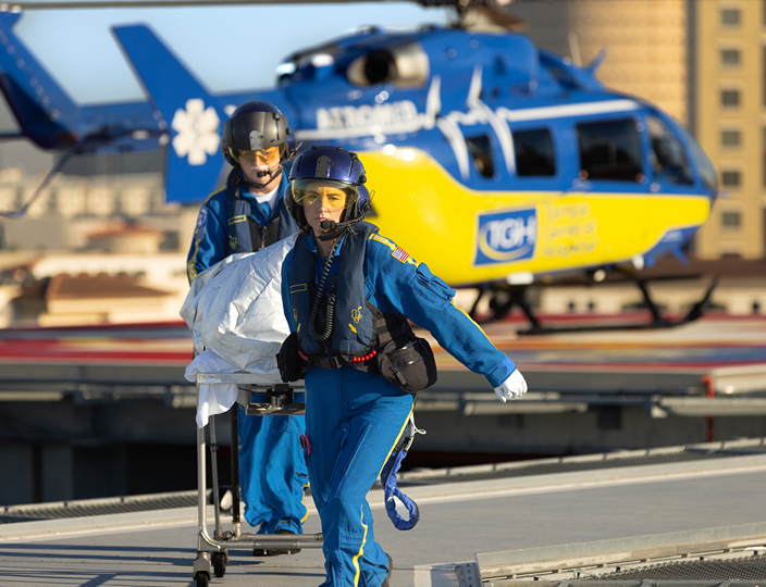 tgh aeromed team members pulling a trauma patient on a covered hospital bed from the helipad to hospital with tgh helicopter and buildings in the background