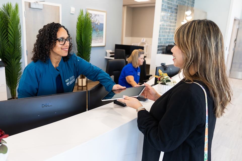 a female patient is handed a tablet by a female staff member while another female staff member works on her computer in the background at the front desk and waiting room area in the tampa general mass general brigham cancer care clinic in palm beach gardens
