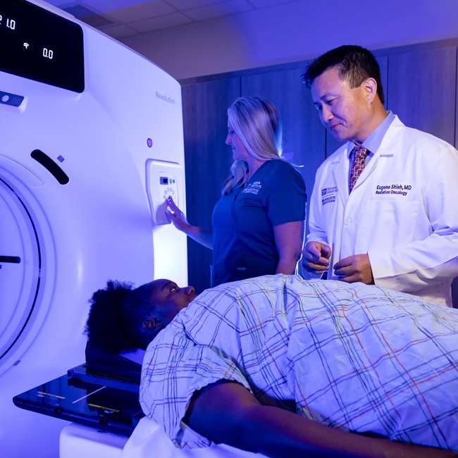 dr. eugene shieh consoling a female patient as she lays on the bed of a ct machine and a ct operator sets up the scan in the background in a dimly lit imaging room
