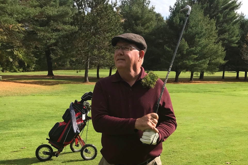 dave colman on the golf course holding a club and smiling after fully recovering from tremors and focused ultrasound procedure