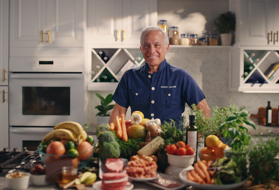 Celebrity chef Geoffrey Zakarian stands behind a kitchen island filled with colorful, plated dishes in a bright, modern white kitchen setting.