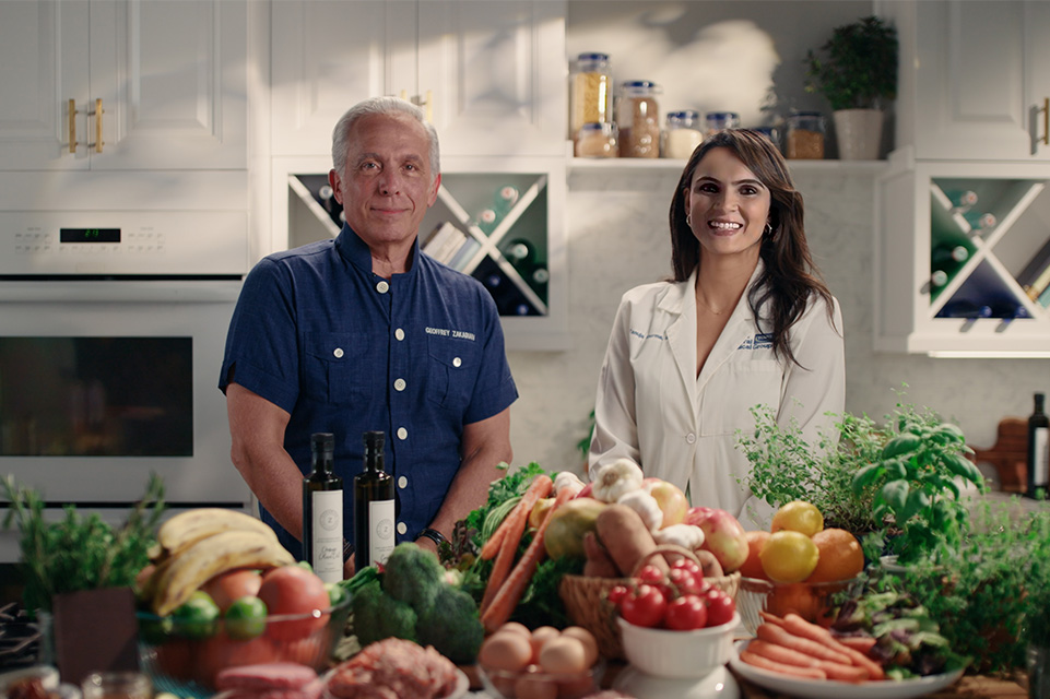 Celebrity chef Geoffrey Zakarian and Dr. Tanuja Sharma stand behind a kitchen island filled with colorful, plated dishes, showcasing Tampa General Hospital’s new patient meals and room service menu in a bright, modern white kitchen