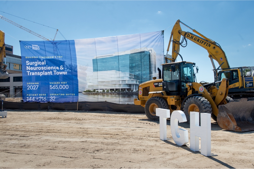 construction vehicles and TGH sign in front of a TGH Taneja Surgical, Neuroscience & Transplant Tower banner at the groundbreaking ceremony
