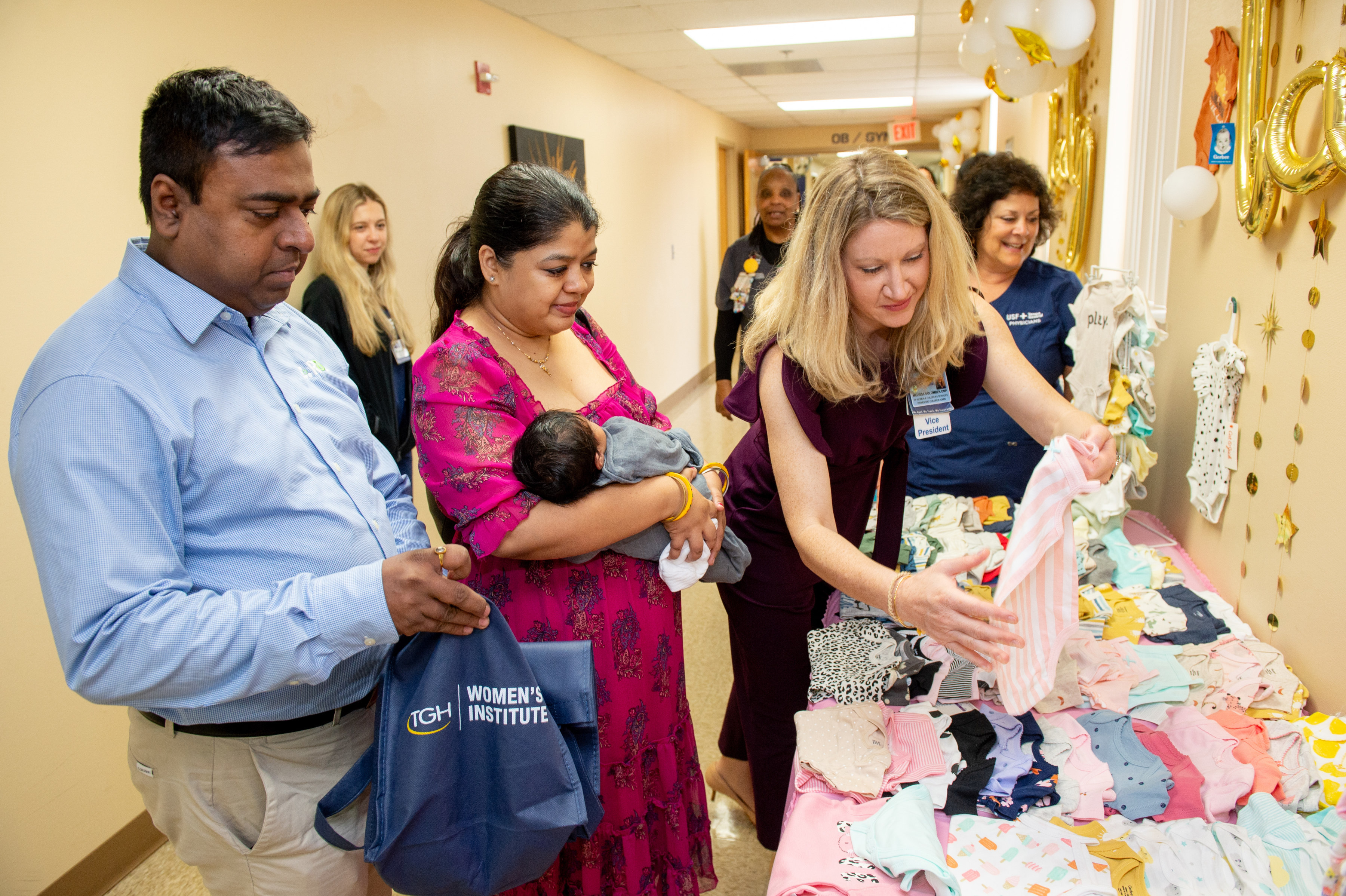 dad and mom holding newborn baby approach a table full of gifts and are handed a onesie by a tgh team member at the genesis baby shower event