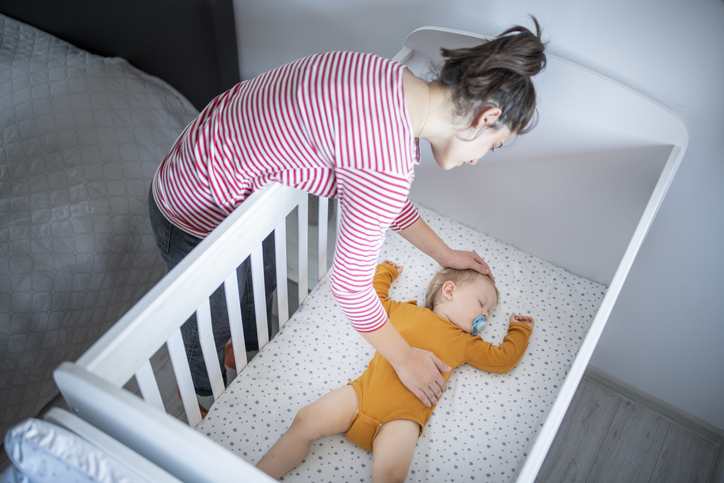 safe sleep guidelines for babies featured image showing mother placing her newborn baby into his crib next to her bed